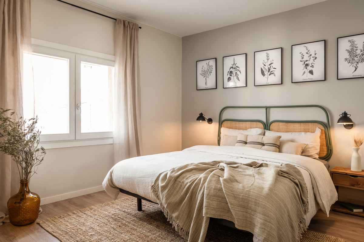 a bedroom with a bed and pictures on the wall. Headboard, linnen bed sheets, big vase on the floor, wall lights, frames above bed with images of herbs. Linnen curtains. Grey walls, beige accessories. Zara home by Aptó interiors.