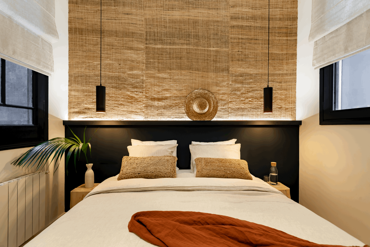 a bed in a bedroom with a beach design. Textured wallpaper, hanging bedside lamps from ceiling, fouling curtains beige linnen. Linnen bedsheets, kentia palm decoration. Terracotta blanket. Interior design, home staging. by Aptó interiors.