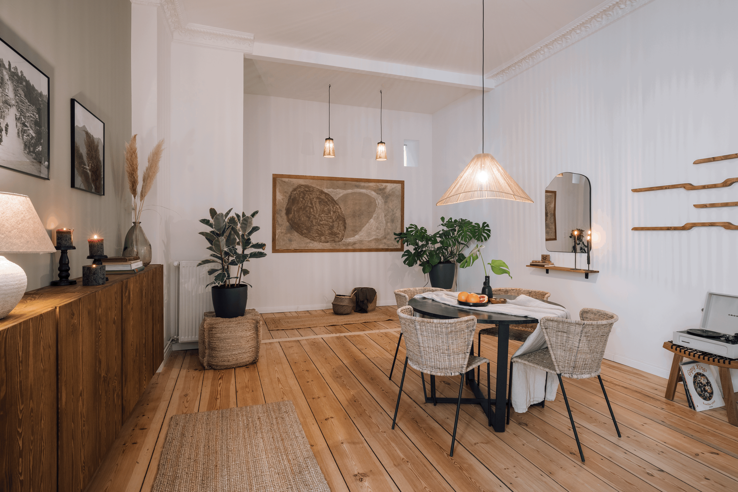 a dining room with wooden floors and white walls. Organic interior design, rattan chairs, organic shaped mirror, big abstract wall art with organic shapes in beige colours. minimalist and cozy dining room setting with roudn table. by Aptó interiors